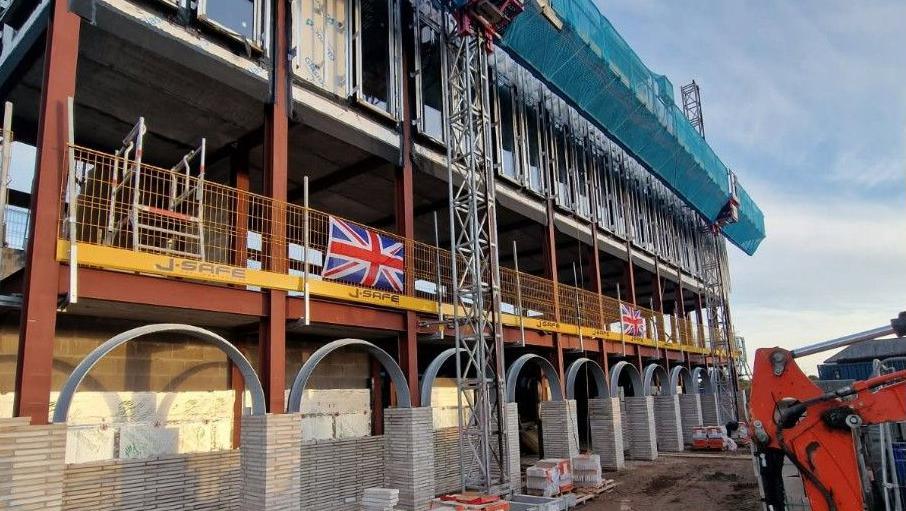 The building site of the mosque under construction in Dalton, Cumbria. The shell of the building is a steel frame, with some windows on the top floors and fencing in the lower levels. Machinery and building materials are around the structure. Two Union Jack flags hang from fencing on the first level.