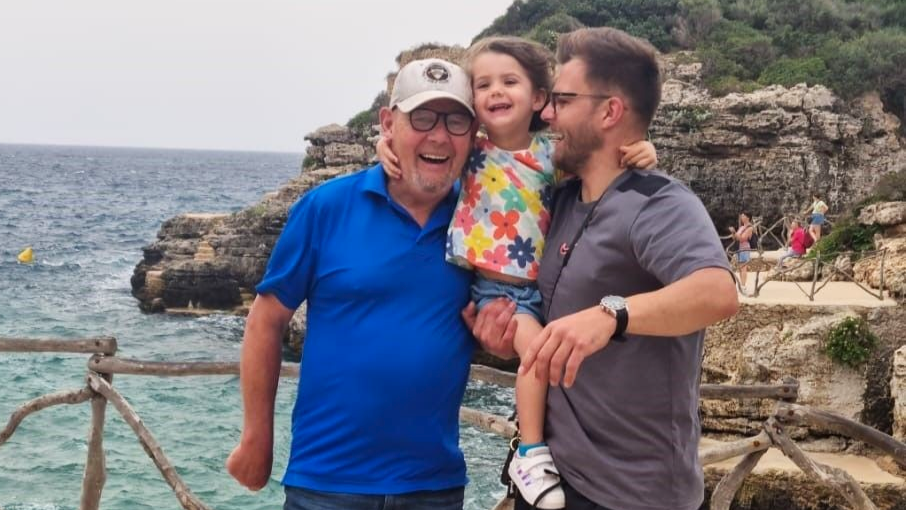 Phillip Marks smiles as he stands on pier with the ocean behind him. He is with his son who is holding his young daughter who has her arm wrapped around Mr Marks' neck.