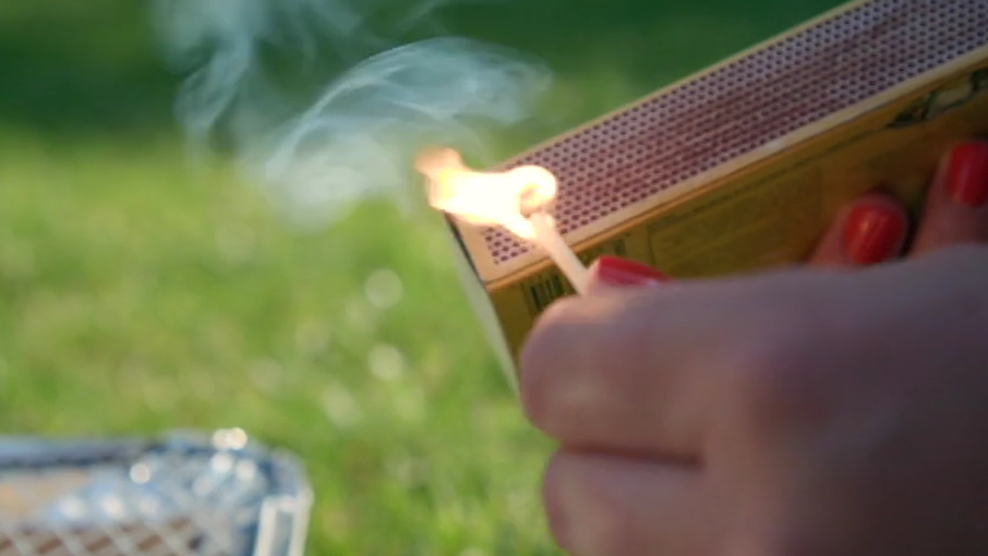 Close up of a hand striking a match against a matchbox. In the background the corner of a disposable barbecue is positioned on some grass.