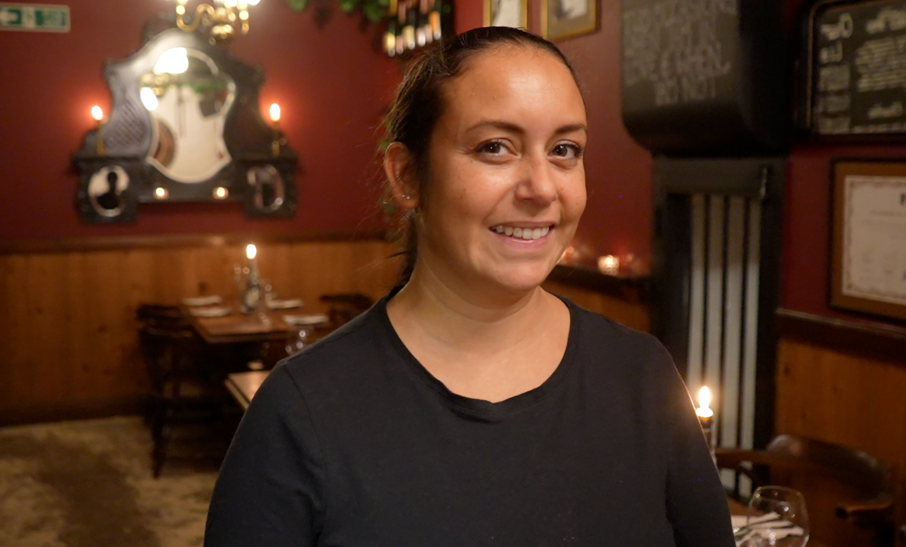 A woman with dark hair which has been tied back smiles at the camera. She is wearing a black top. The walls behind her are a rich red colour and candles are on the dining tables.