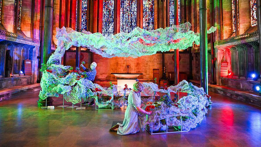 A floral display with lots of material shaped to look as though it is floating in a cathedral with dramatic coloured lighting. Two people making finishing touches with flowers.