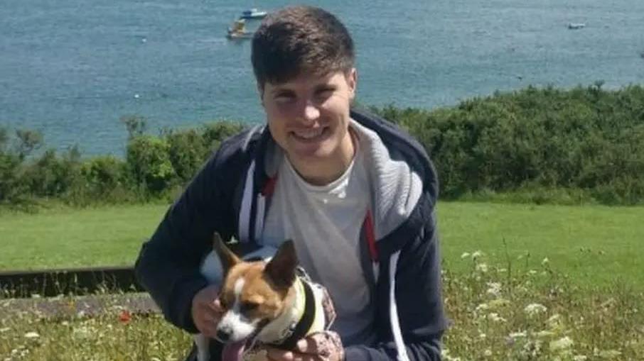 Adam Ellison, with brown hair wearing a blue hoodie and grey t-shirt, cuddles his dog on the river bank. He is smiling.