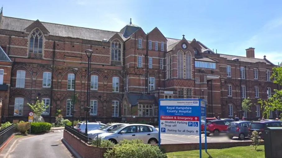 Winchester Hospital, red brick building with car park in front and a NHS blue and white sign for directions to the building.