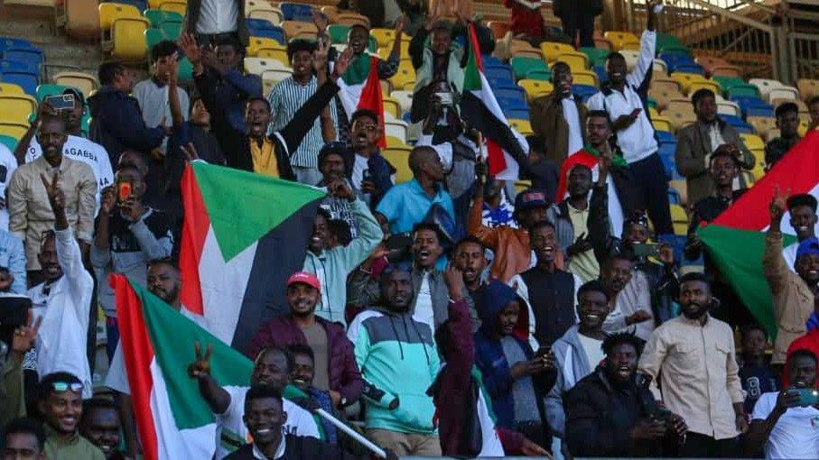 A group of around 30 male Sudan fans smile and wave their arms and flags to celebrate qualifying for Afcon 2025. They are clustered towards the bottom of the image, with empty blue, green and yellow seats higher up in the stand