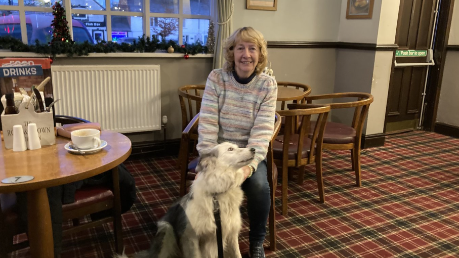 A woman in a light striped jumper and jeans sits in a chair and smiles at the camera. A large white and grey dog sits at her feet. She is sat in a pub, with tables and chairs around her, with a cup, cutlery box and a menu on the table to her left.