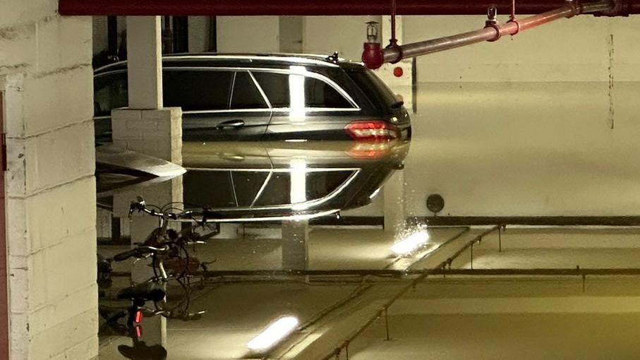 A flooded car park with the water level up to the break lights of two parked cars in the background. The rear brake lights are reflected in the water