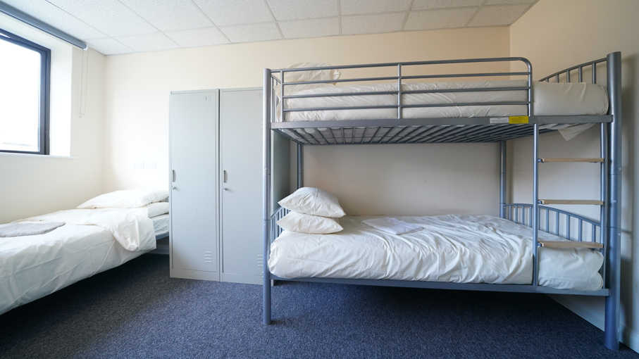 Room with bunk beds. The carpet is blue. The bunks and single beds have metal frames and white sheets. There are three beds in the room.