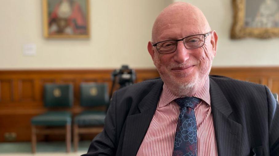 A man with a white beard wearing glasses, a striped red and white shirt, patterned tie and jacket sitting in a council house room