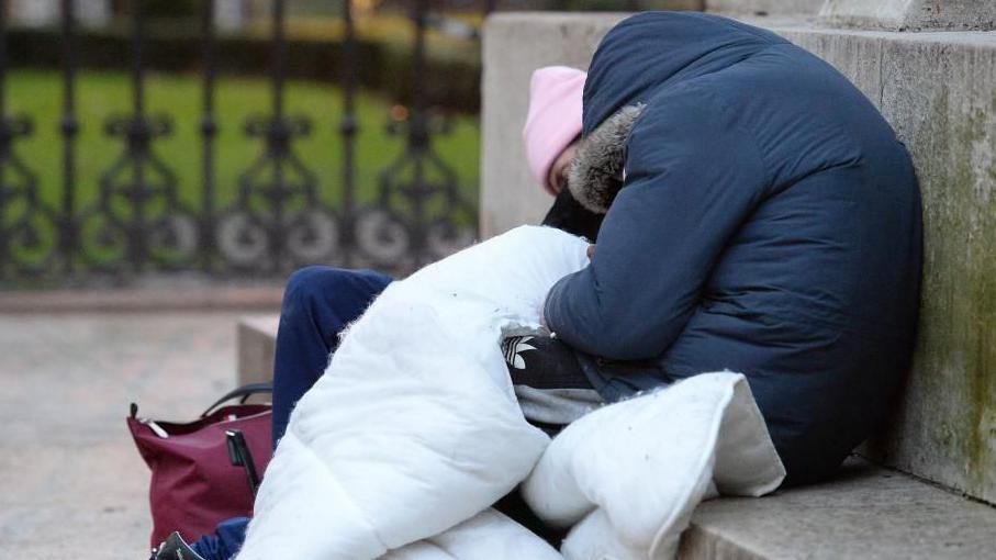 Two people facing away from the camera sitting on a concrete step on the street. The person closest to the camera is wrapped in a white duvet and is wearing a navy blue winter jacket with the hood up. The person behind them has a red bag and is wearing a pink hat. 