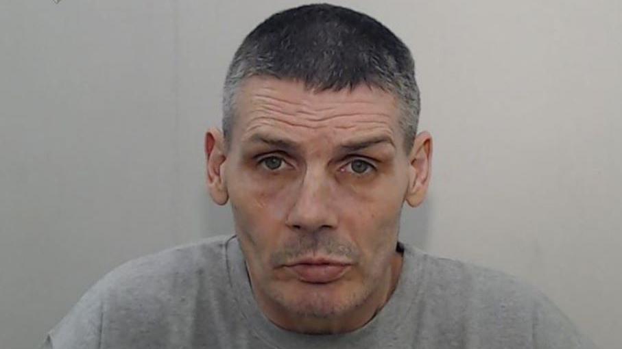 Police mugshot of Peter Pitt. He has very short grey hear and is wearing a light grey t-shirt.