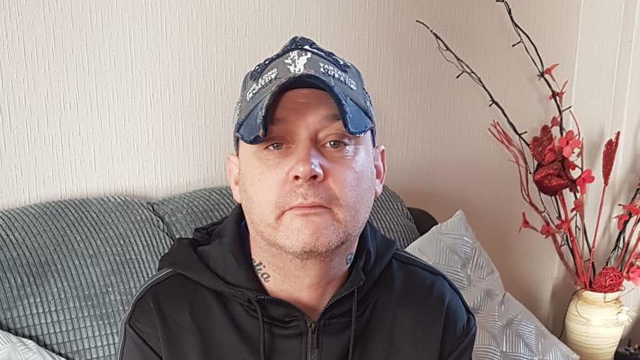 A man with a grey cap and black hoody sitting on a grey sofa with red flowers on the right. He has brown eyes and tattoos on his neck.
