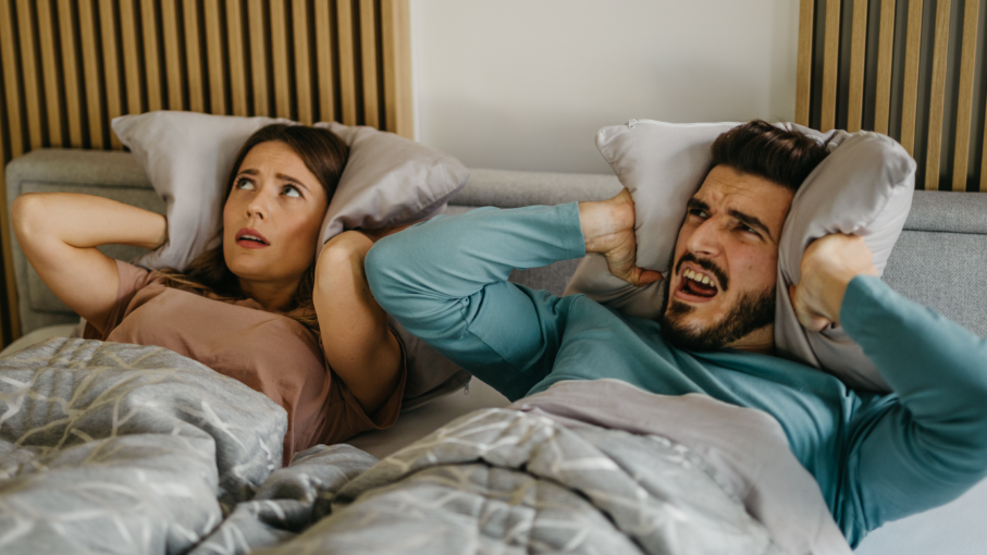 A couple  suffering from noisy neighbours lie awake in bed. A woman clamps a pillow over her ears while her partner has his mouth opened angrily as he looks at the ceiling.