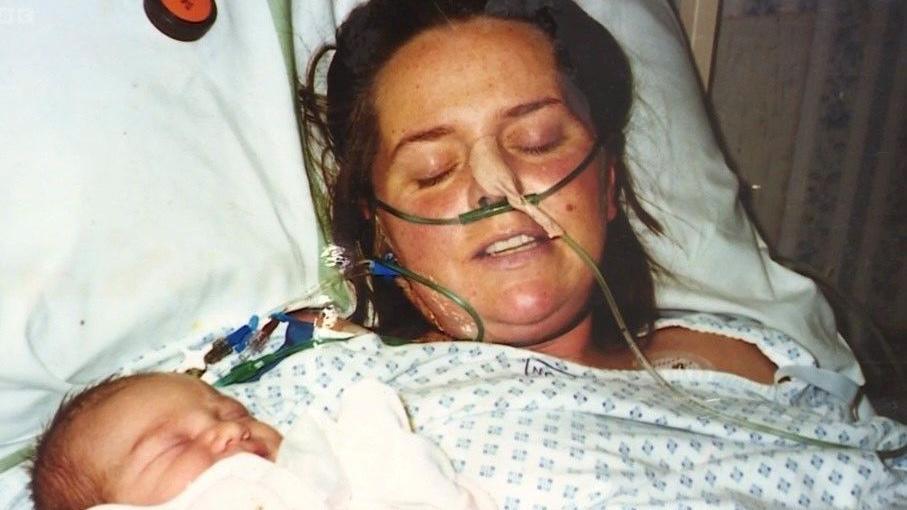 Anna Edwards next to her baby Somer in hospital. She is unconscious and has a tube in her nose. The baby has some dark hair and is wrapped in a white blanket asleep.