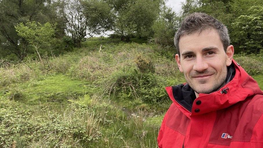 Dr Chris Hatcher stands in the countryside, smiling in a red coat. The ground behind him is boggy and rugged, with plants growing on the ground and trees in the background.