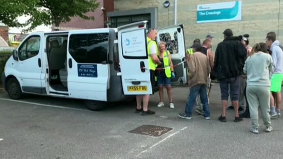 Men and women queue to receive food and drink from the back of a white van, which has "Help the Homelezz" signs on the doors. Two volunteers wearing yellow hi-vis jackets are handing out donations.