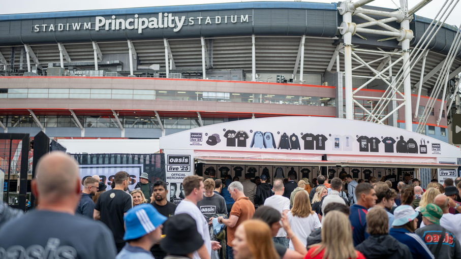 Fans queuing at an Oasis merchandise pop up stand with the Principality Stadium in Cardiff seen in the background. At the top of the stand is a white roof showing the different items for sale, including bucket hats and black t-shirts and jackets.