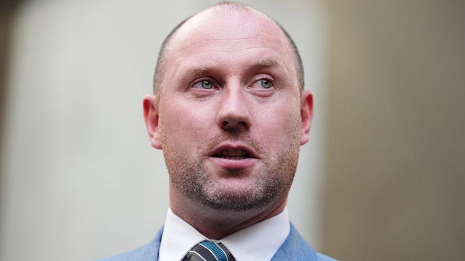 Neil Gray, a bald man in a white shirt and blue suit, looks to his left