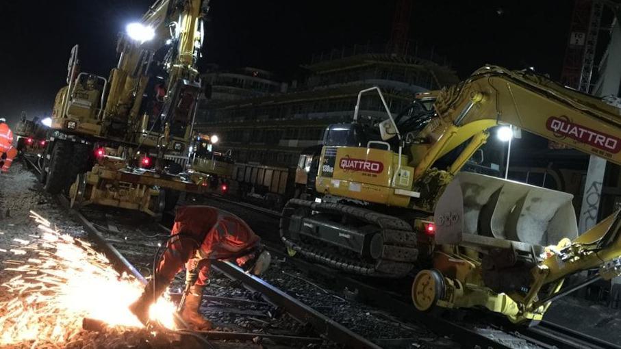 It is dark, with large pieces of machinery on rail lines and a person working with a welding tool on a piece of track.