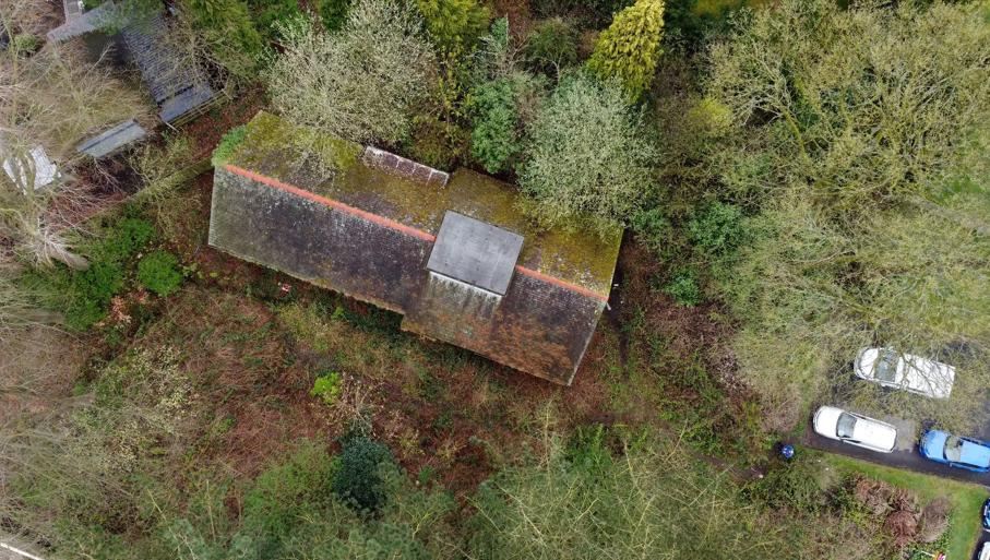 An aerial view of a large tiled roof building. There are cars to the right of the picture, and the building is surrounded by trees and bushes 