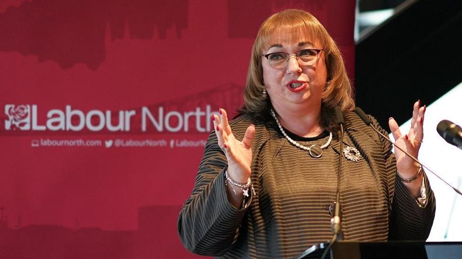 Sharon Hodgson talking in front of a microphone. She has short, brown hair with a full fringe and square glasses. She is wearing red lipstick and a pearl necklace. She is talking with her hands and is standing in front of a red Labour North banner.