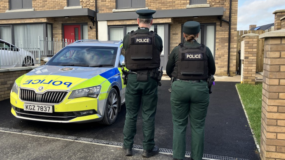 Show a police car in yellow, light green blue and grey with the PSNI cresh at the word 'police' on the bonnet. Two police officers, a male and female, in green uniforms and black flak jackets with the word 'police' stand to the right of the car in the driveway of a house, facing the house. 