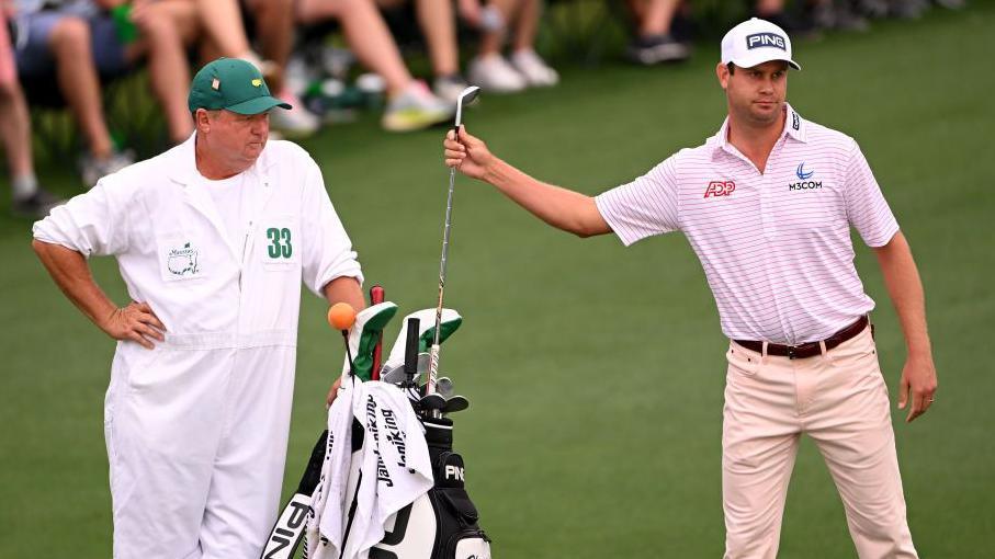 Harris English is watched by caddie Eric Larson as he holds a club by his golf bag at the 2023 Masters