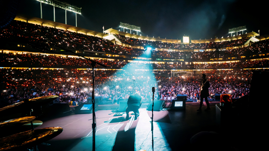 An image taken from the back of the stage as My Chemical Romance are performing. Gerard Way is in the centre, kneeling on the floor and facing the audience of thousands of people. He is under a spotlight, and his head is bowed forward. The stadium is lit up as it is nighttime.