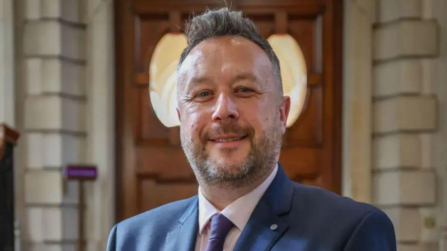 A man wearing a suit smiles at the camera. He has short, brushed-back gark hair, and a slightly greying beard. 