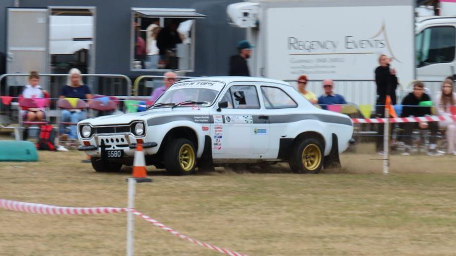 A white classic rally car is driven around the grass at the showground during the North Show in Guernsey. Several people are sitting outside the arena watching the car drive past.