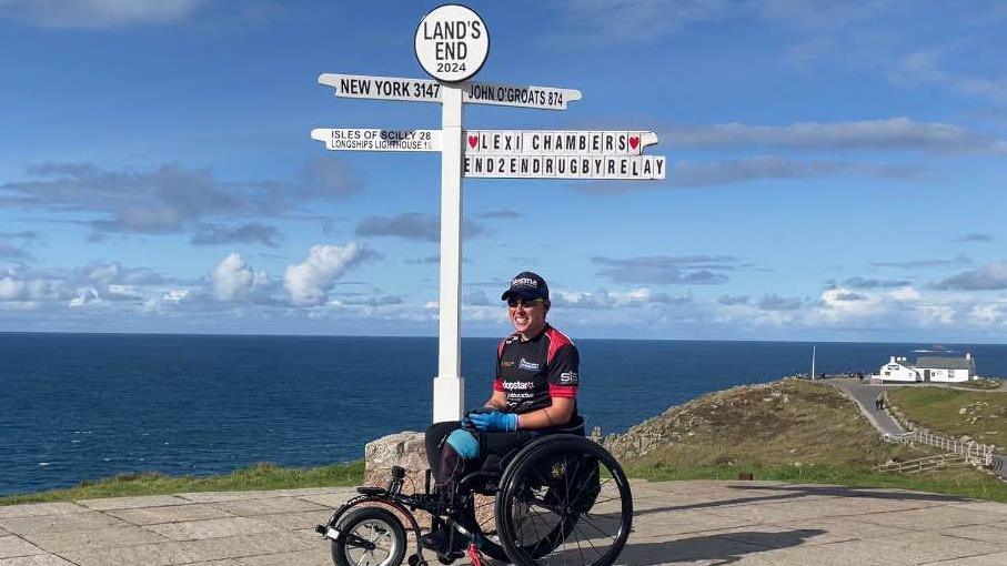 Ms Chambers, wearing a black and read sports shirt, in her wheelchair. Behind her is the lands end sign pointing at New York, John O'Groats and Isles of Scilly. There is one sign that says Lexi Chambers END2ENDRUGBYRELAY on it. The background is the sea. In the left of the background is a hill with a white house. 