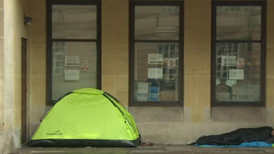 A rough sleeper lying near a tent beside a building. He is wrapped in a blanket. The ground is wet.