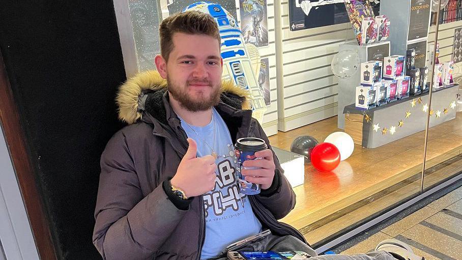 A man wearing a coat sits on a chair making a thumbs up sign with one hand and holding a coffee cup in the other