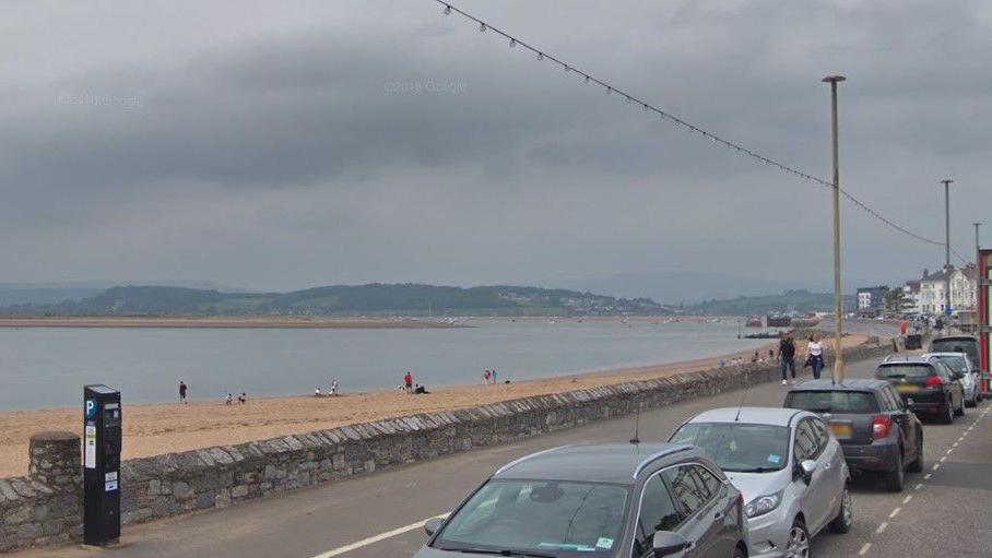 Image shows Exmouth seafront on a grey day. A number of cars are parked in the bays on the left-hand side of the image. People are walking along the esplanade behind the beach. There is a stone wall separating the walkway and the sandy beach. A number of people are standing on the sand. 