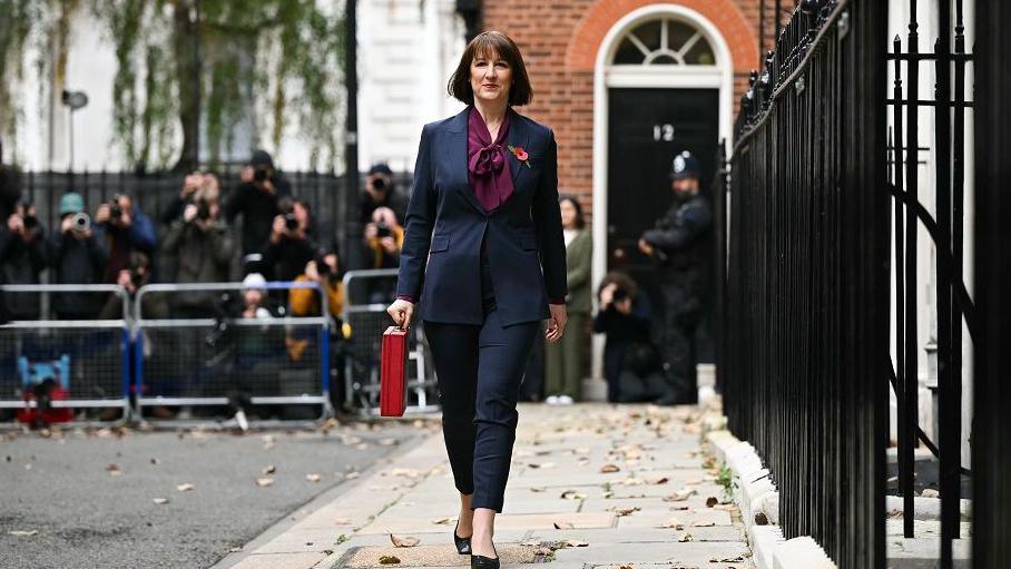 Rachel Reeves carries the red Budget Box as she leaves 11 Downing Street. A crowd of journalists and camera operators are in the background, standing behind a fence.