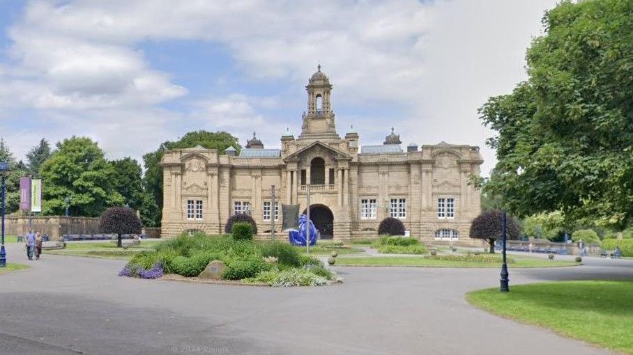 A grand stone building with a central tower, with a path and greenery in the foreground and a tree to the right-hand side