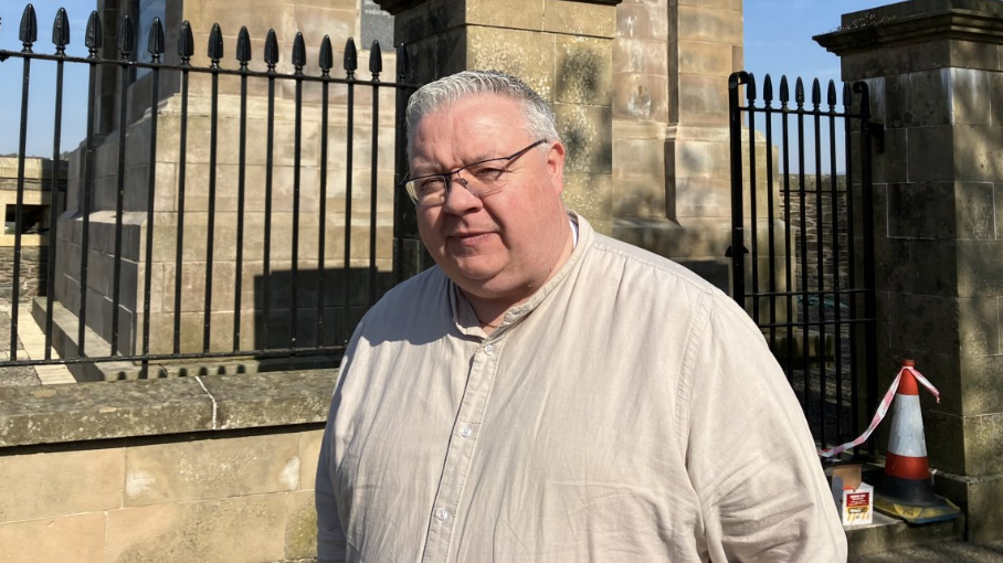Shows a man in a light coloured shirt with glasses and grey hair standing in front of railings 