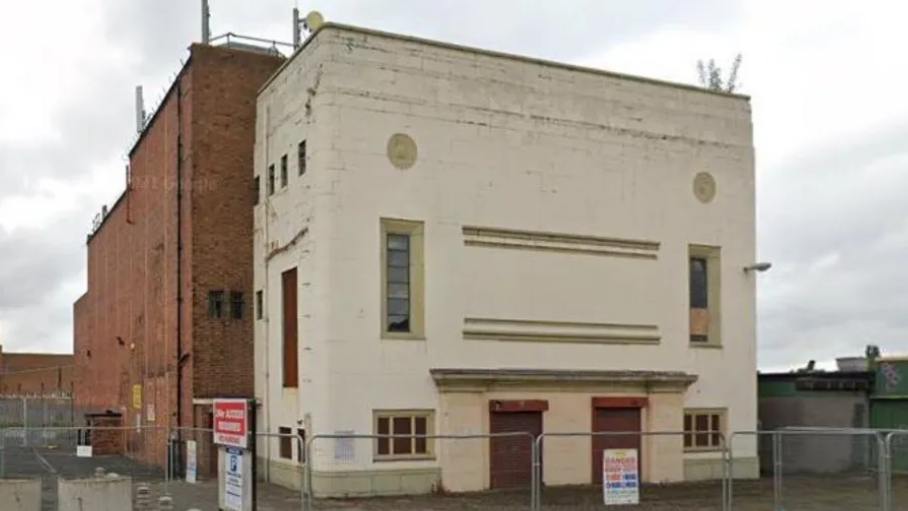 A slab-fronted multi-storey cream coloured building, with red brick walls to the side, surrounded by security fencing 