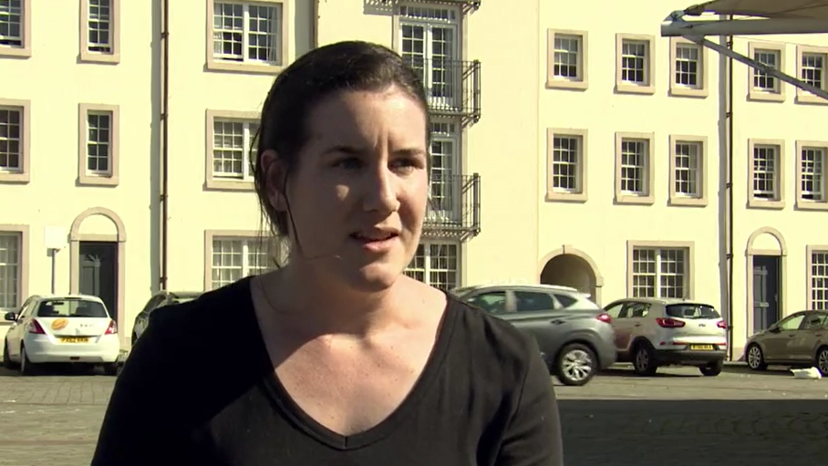 Hannah Denny in front of a yellowy-white, well-maintained terrace building. She has dark hair and is wearing a black top. 