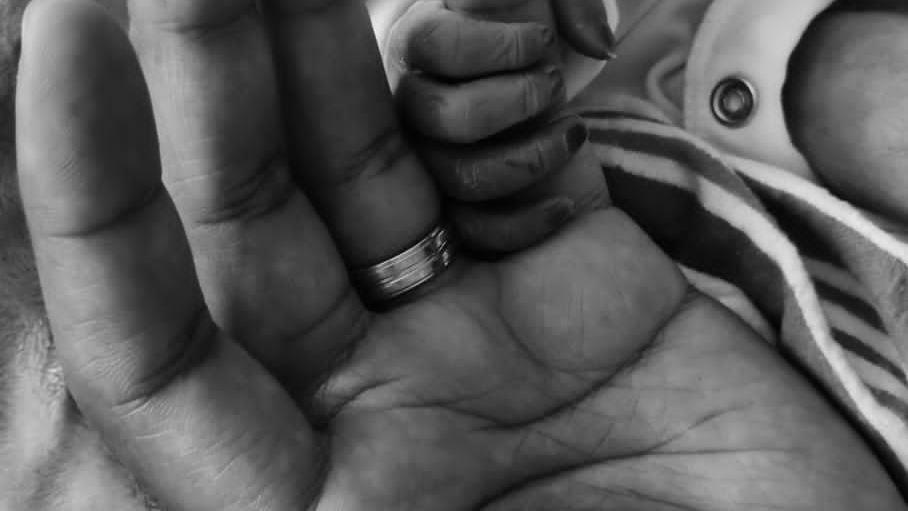 A black and white photo of an adult hand, with a ring around the third finger, with the little finger being grasped by a much smaller hand