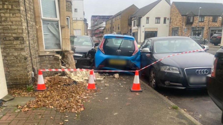 The rear of a blue car is seen wedged between two parked cars. To the left is a house with part of its lower wall destroyed by the car that crashed into it. There are police cones and tape cording off the scene. 