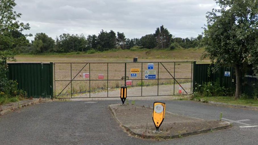 The former Festival Gardens entrance closed off with a fence with dry grass behind and junction with two yellow faded traffic bollards.
