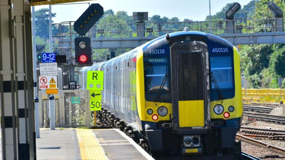 A blue South Western Railway train arriving alongside a train platform, displaying the destination 'Alton' on the front