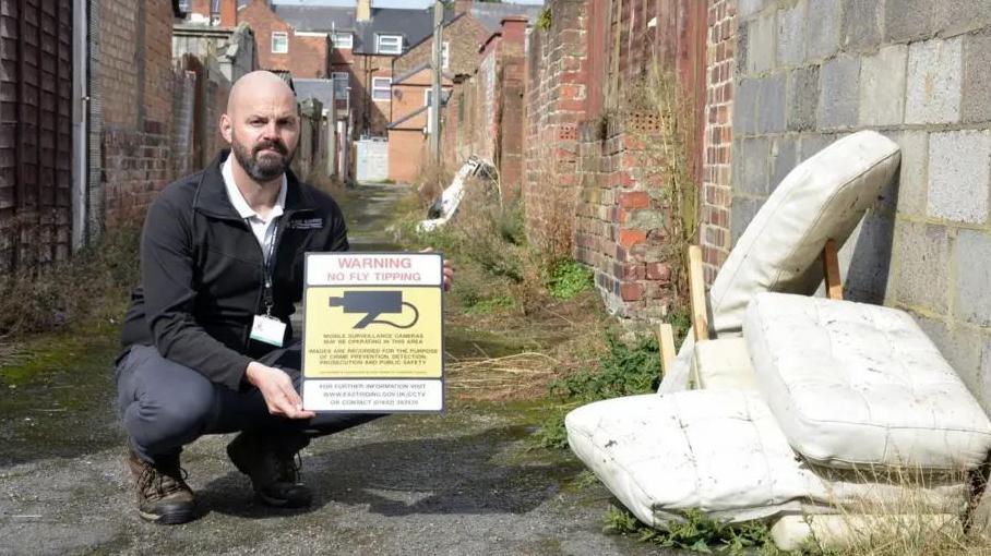 A man holds a 'warning - no fly tipping' poster with a CCTV camera on it, he poses next to a fly-tipped sofa in a back alley. 