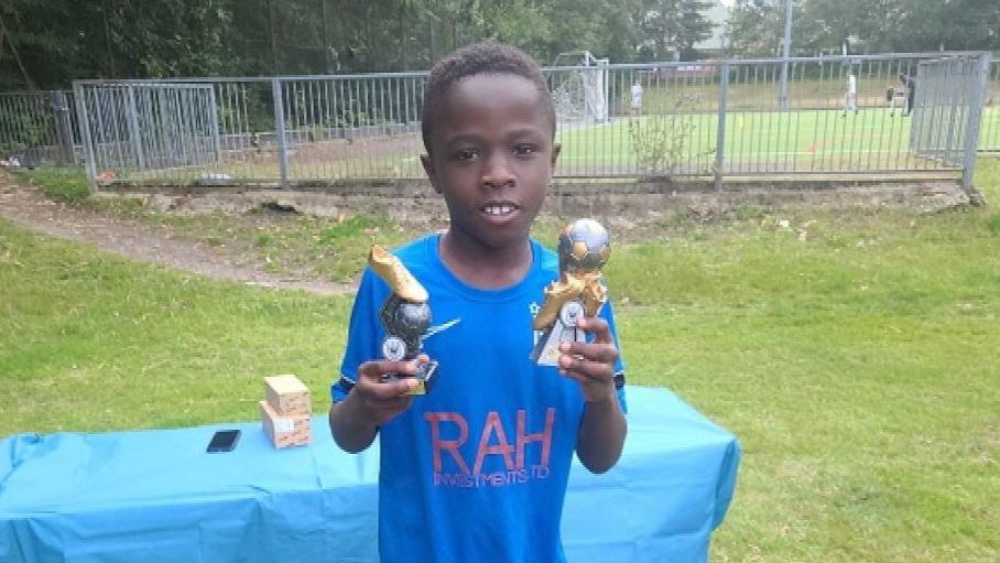 Othneil Adoma wearing a blue football kit holding two trophies in a park
