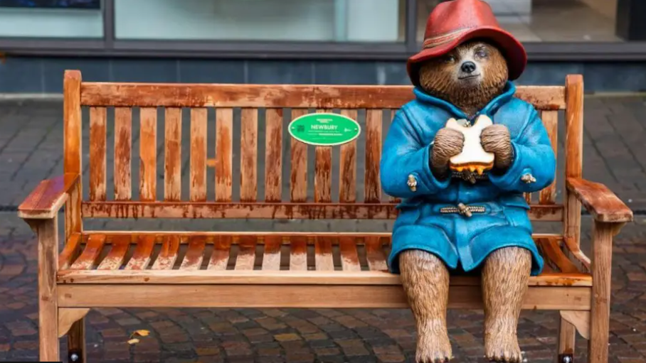 A statue of a Paddington Bear in a red hat and a blue coat holding a sandwich and sitting on a bench