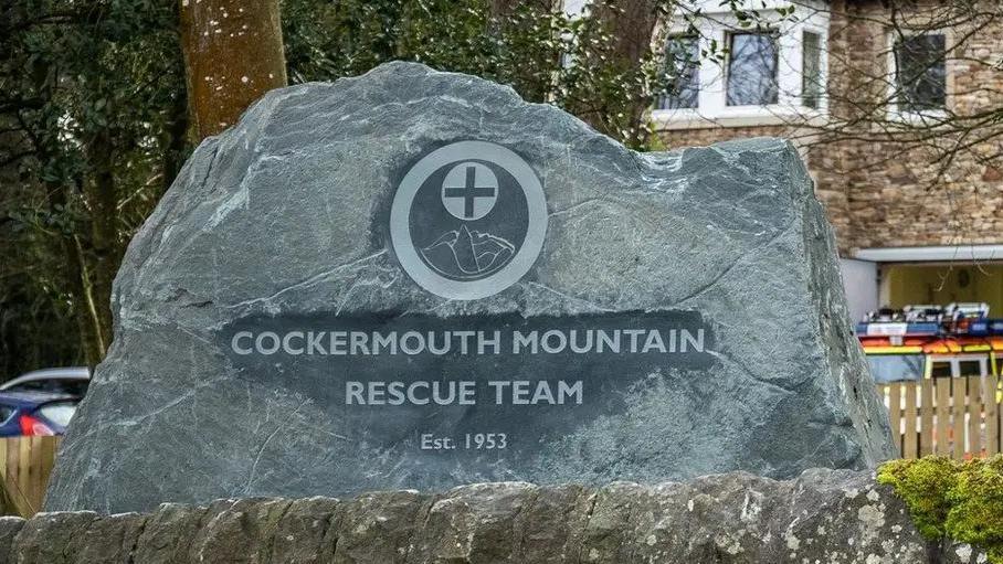 A slate sign which has 'COCKERMOUTH MOUNTAIN RESCUE TEAM' engraved on it. The sign is placed in front of the mountain rescue team's headquarters.