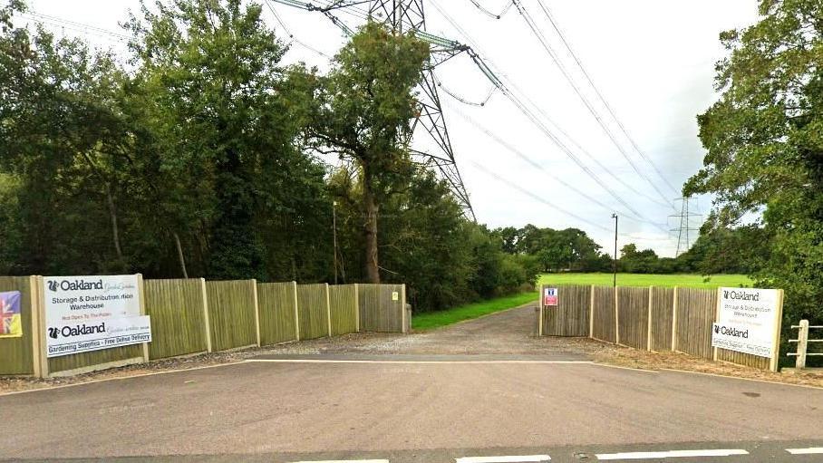 The entrance to the site owned by the company. We can see signs for Oakland Gardening Supplies, fencing and an entrance of a driveway that leads to a field.