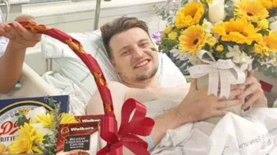 A man with brown hair and a small beard in a white hospital bed holding a small gift with yellow flowers behind him and a hand holding a basket of food with a red bow on it.