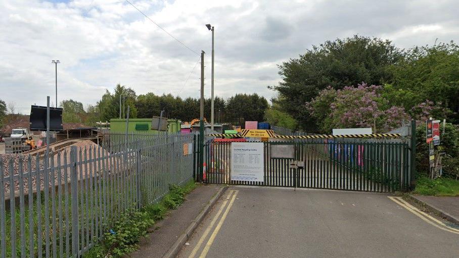 A set of green gates with black and yellow bar across the top at the entrance to the tip with a sign detailing the site's opening hours. There are trees, vehicles and storage containers visible in the background.