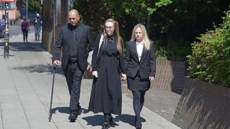 The family of Elise Sebastian walking towards the camera, which, left to right, includes her father, auntie and mother. They are all dressed in black and are walking along a pavement. The road is to their right and a brick wall and bushes are to their left. It is a sunny day.